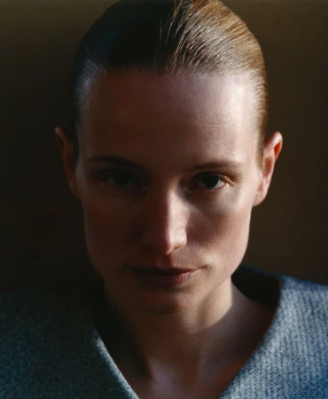 Close-up portrait of an adult woman with slicked-back short hair, wearing a grey top, set against a dark, gradient background.