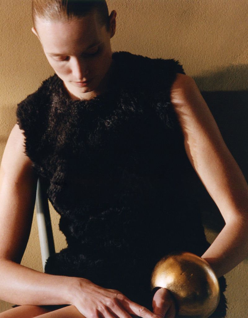 A stylish adult female with fair skin sits on a chair with a gold bracelet, wearing a dark vest and looking downward in an indoor setting.