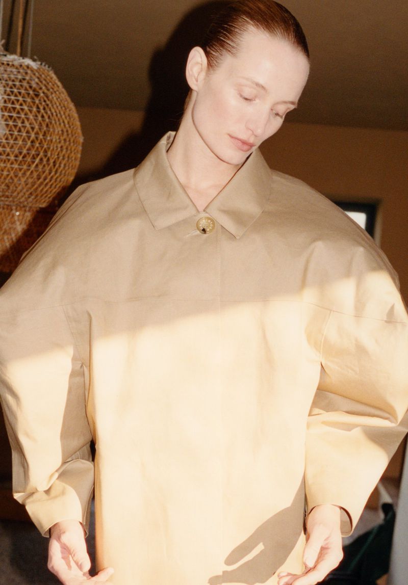Fashion shot of a fair-skinned woman in a neutral overcoat standing in an indoor environment with natural light and a calm expression.