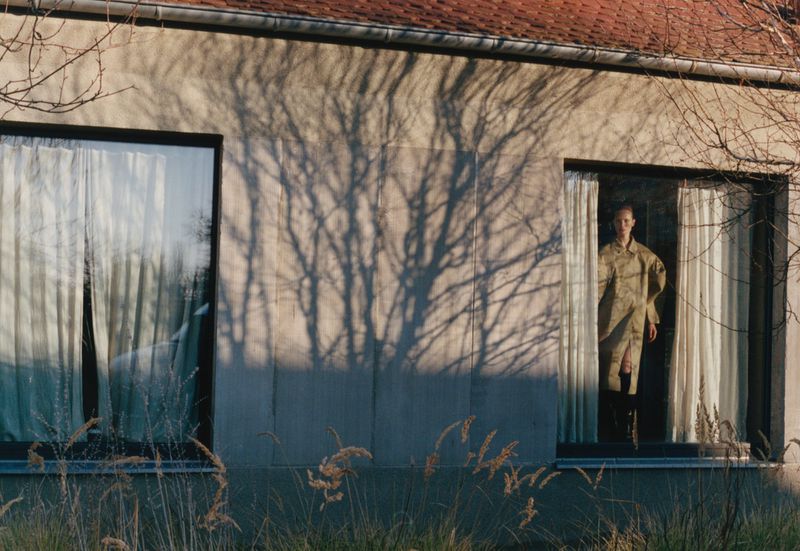 Woman in trench coat stands in window of sunlit house with a tree shadow on the wall, long golden grass in foreground.