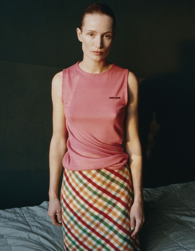 A woman poses indoors, wearing a pink tank top and plaid skirt, against a neutral background, with soft, natural lighting. Fashion portrait.