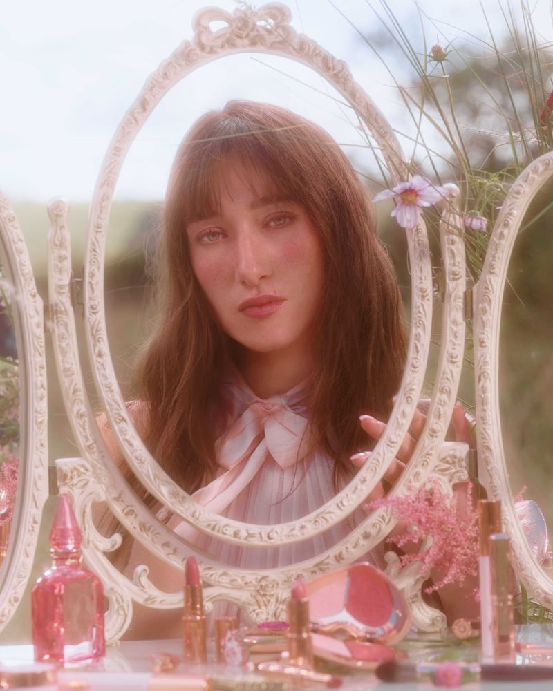 Woman with bangs and long brown hair looking at her reflection in a vanity mirror surrounded by makeup and pink flowers.