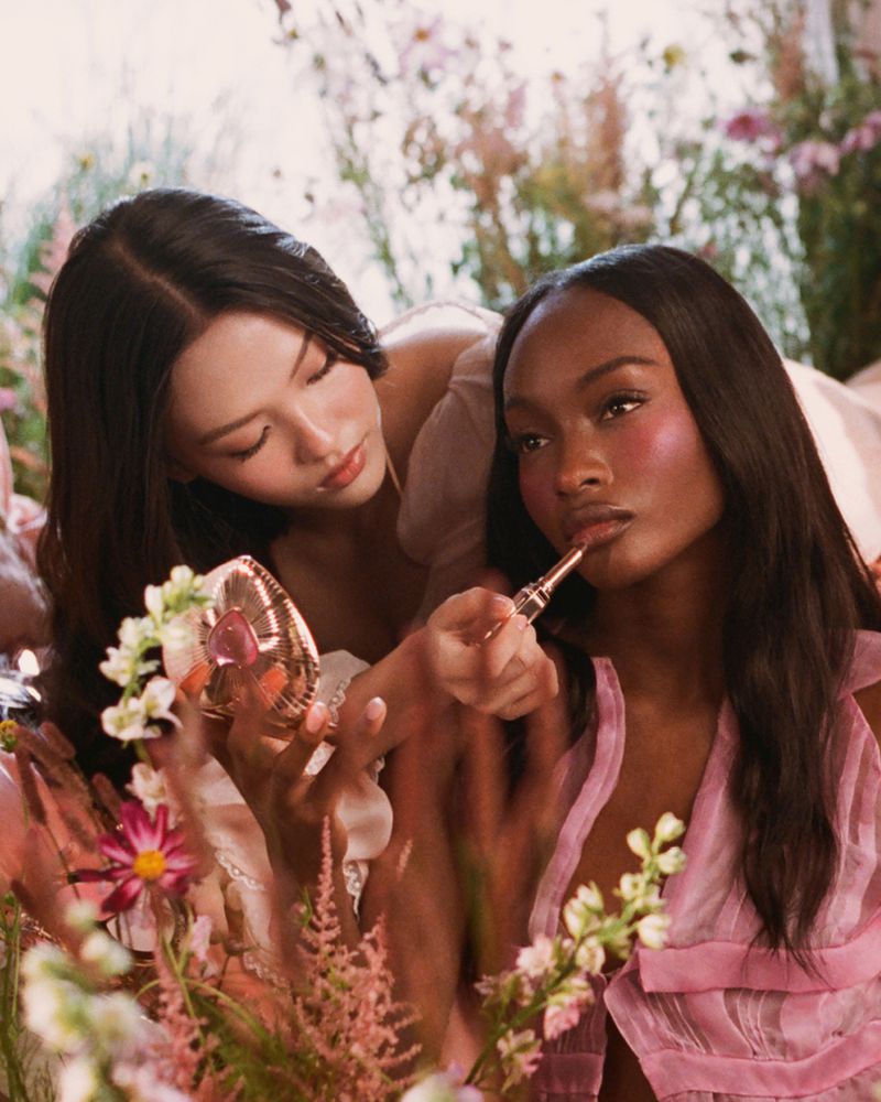Two women apply makeup in a floral garden setting, one holding a compact mirror and the other applying lipstick, both wearing pink tops.