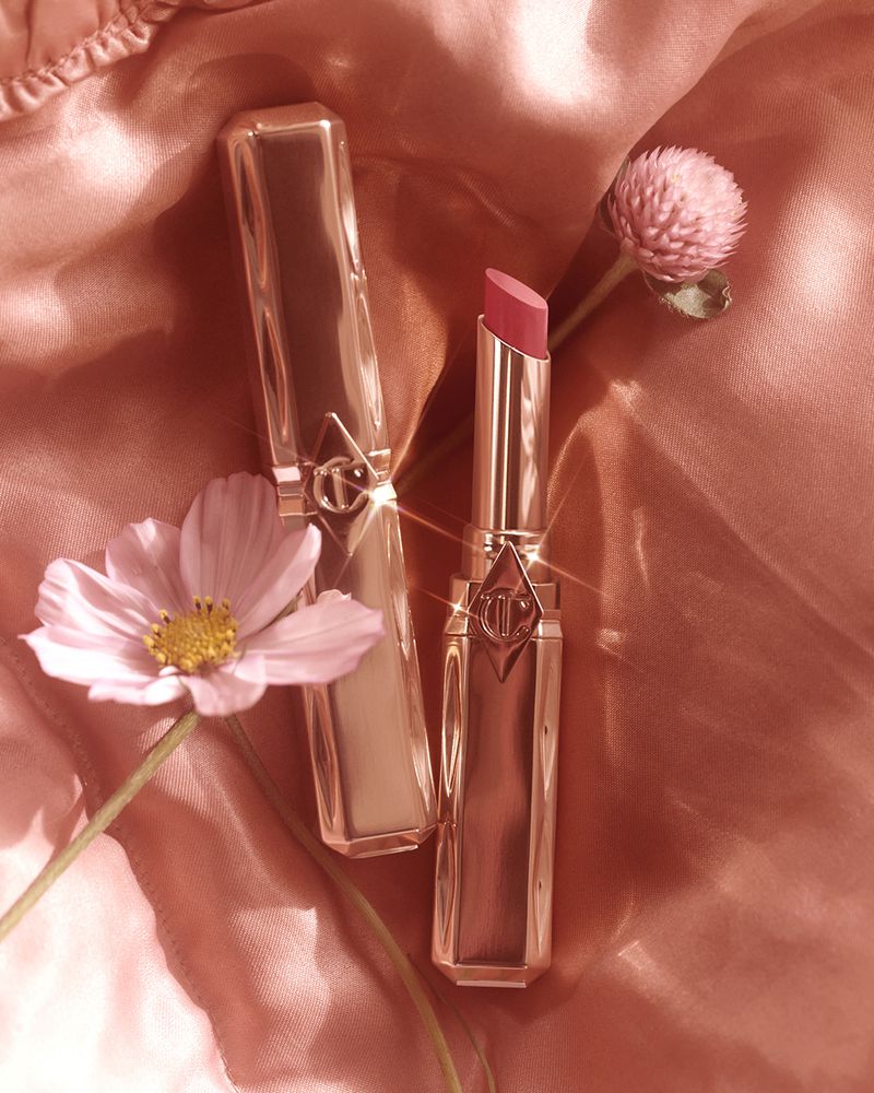 Two gold lipstick tubes, one open with pink lipstick, arranged with pink flowers on a shiny pink fabric backdrop in a studio shot.
