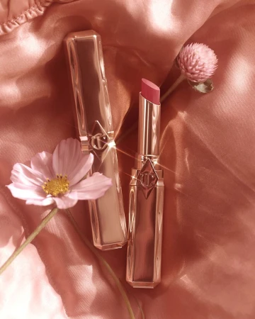 Two gold lipstick tubes, one open with pink lipstick, arranged with pink flowers on a shiny pink fabric backdrop in a studio shot.