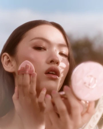 Close-up of a woman applying light pink powder to her face using a cosmetic puff outdoors against a soft blue sky background.