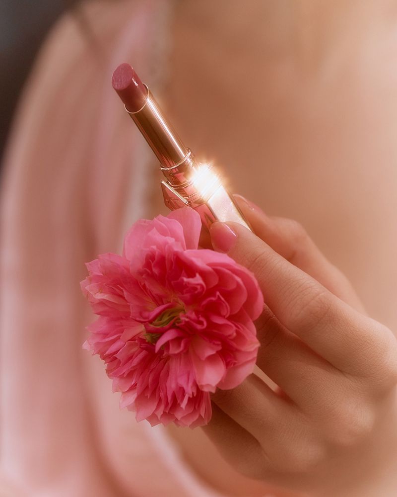 Close-up of a woman's hand holding a pink lipstick tube and a pink flower against a soft, blurred background, beauty and cosmetics.