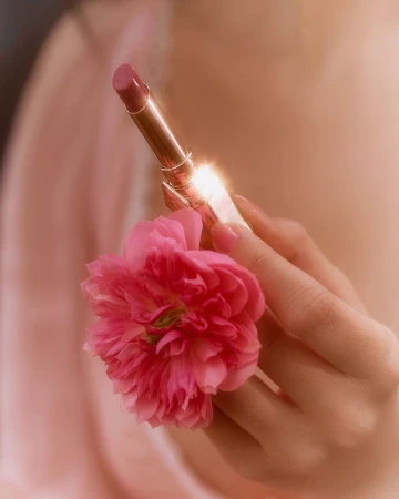Close-up of a woman's hand holding a pink lipstick tube and a pink flower against a soft, blurred background, beauty and cosmetics.