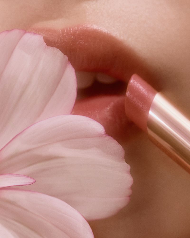 Close-up of a woman applying pink lipstick with a flower detail against a soft, blurred pink background for beauty marketing.