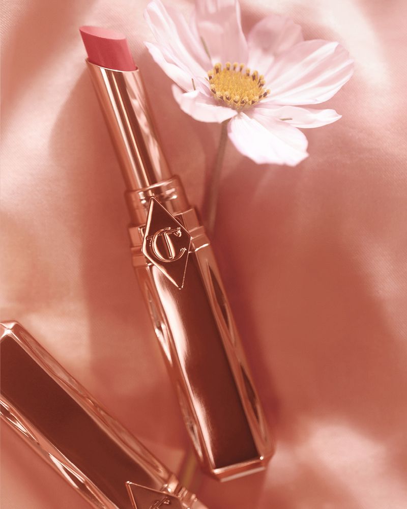 Close-up of a pink lipstick tube by Charlotte Tilbury next to a white flower against a pink satin background. Beauty and cosmetics.