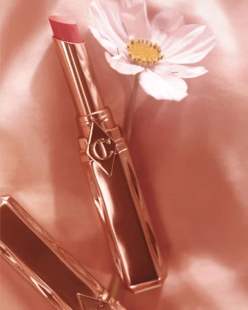 Close-up of a pink lipstick tube by Charlotte Tilbury next to a white flower against a pink satin background. Beauty and cosmetics.