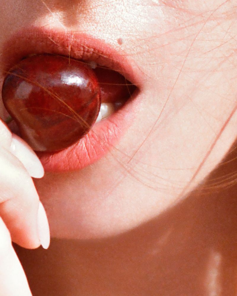 Close-up of a woman's mouth with light pink lipstick and a dark red cherry held in her teeth, showcasing beauty and food.