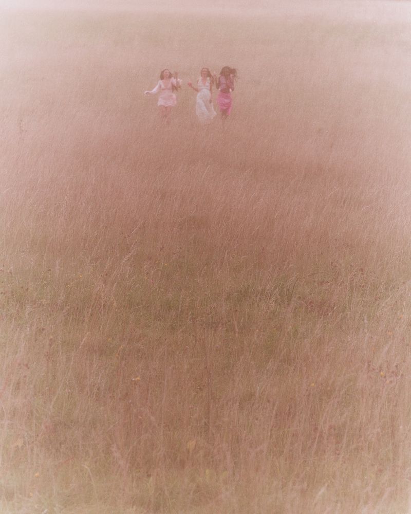 A group of young women are running hand-in-hand through a tall grass field on a hazy day, creating a dreamy, carefree atmosphere.