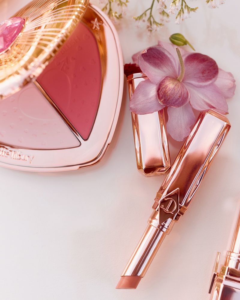 Rose gold makeup set, including blush compact, lipstick, and applicator brush, against a light backdrop with floral accents. Beauty product shot.