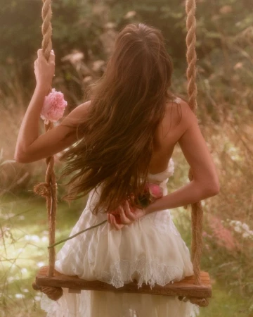 Back view of a woman in a white dress sitting on a swing, holding pink roses against a blurred background of green foliage.