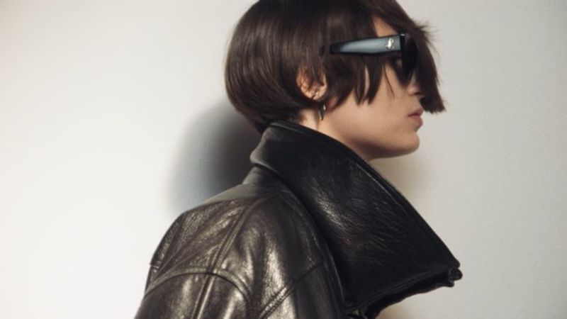 Side profile of a brunette adult wearing a black leather jacket and dark sunglasses against a light background in an indoor setting.