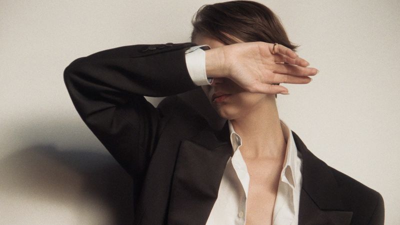 Elegant portrait of a stylish woman in a black suit and white shirt, covering her face with her hand against a neutral backdrop.