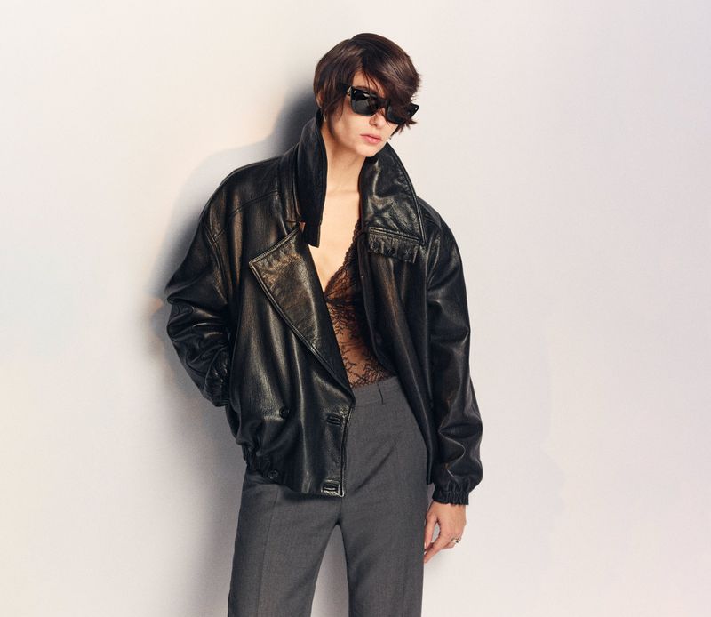 Adult female wearing a leather jacket, lace top, and grey pants poses in a medium shot against a neutral backdrop in a fashion setting.