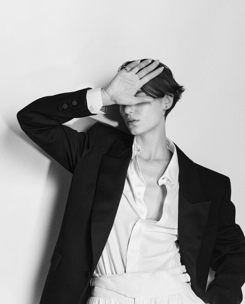 Monochrome portrait of a young adult in an unbuttoned shirt and jacket with a hand covering their eyes against a white background.