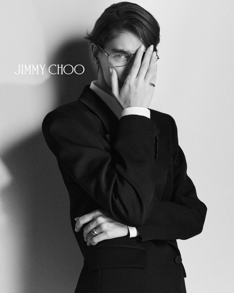Monochromatic fashion portrait of a woman with short hair and eyeglasses wearing a suit, her face partially obscured by her hand.