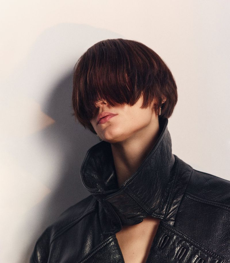 Close-up studio portrait of an adult woman with short brunette hair covering her face, wearing a black leather jacket.