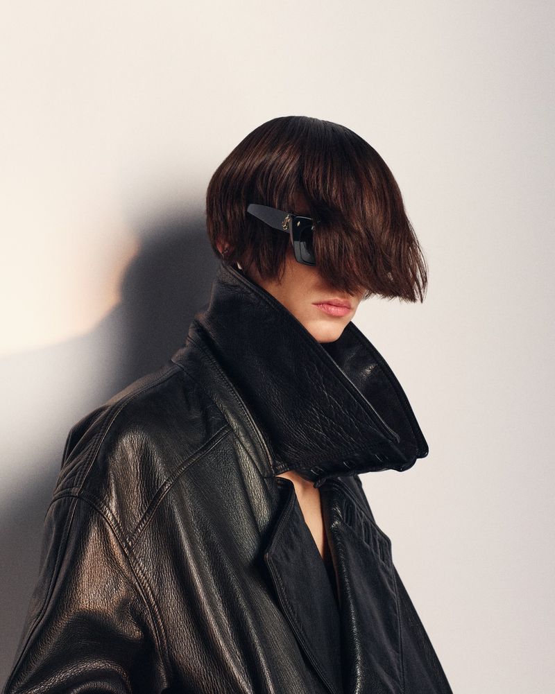 Person with dark hair and sunglasses in studio wearing black leather jacket against off-white background in a fashion editorial style.