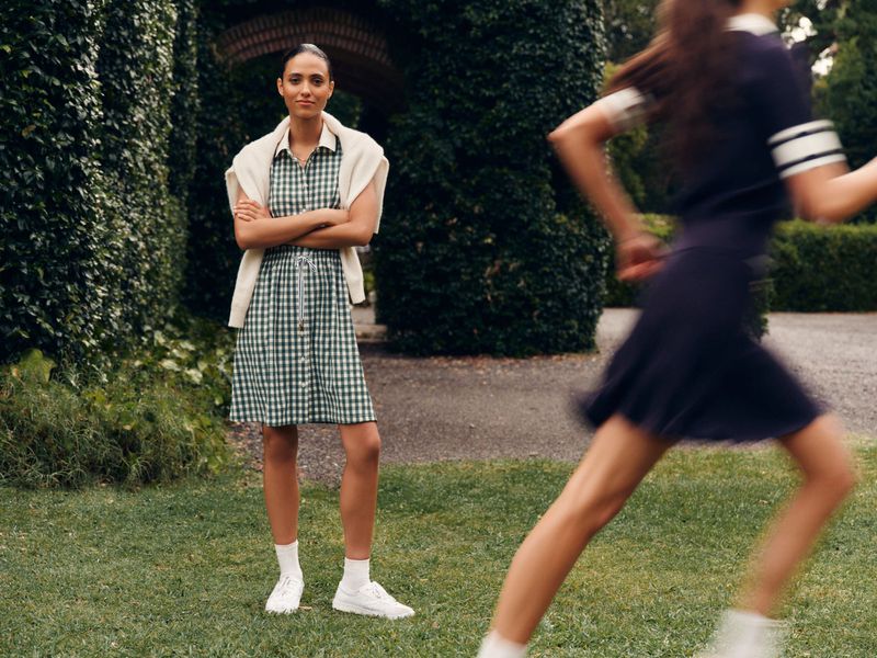 Woman in a green gingham dress standing on grass while a second woman runs in the background on a green, outdoor location.