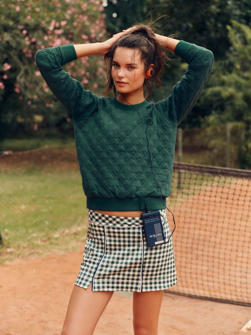 Woman in a green quilted sweatshirt and a checkered miniskirt with a portable cassette player on a tennis court in daylight.