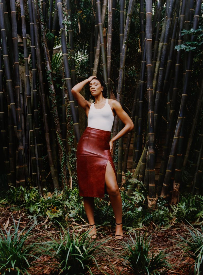 Woman in a sleeveless white top and red skirt poses in a wooded area with tall bamboo stalks in the background, fashionable and elegant.