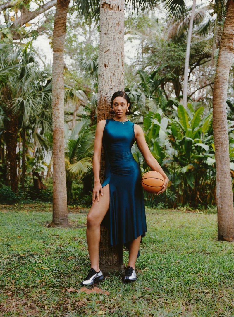 A woman poses in a teal dress and sneakers, holding a basketball in a lush outdoor setting with palm trees and dappled sunlight.