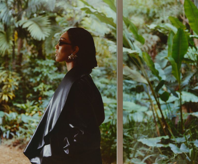 Woman in a black coat and sunglasses stands against a backdrop of a tropical forest with lush green plants and diffused light.