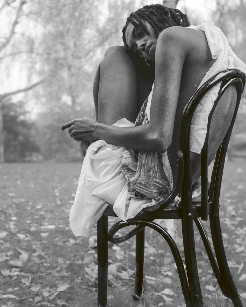 Black and white portrait of a slender person with dreadlocks, sitting on a wooden chair outside in a grassy area with trees.