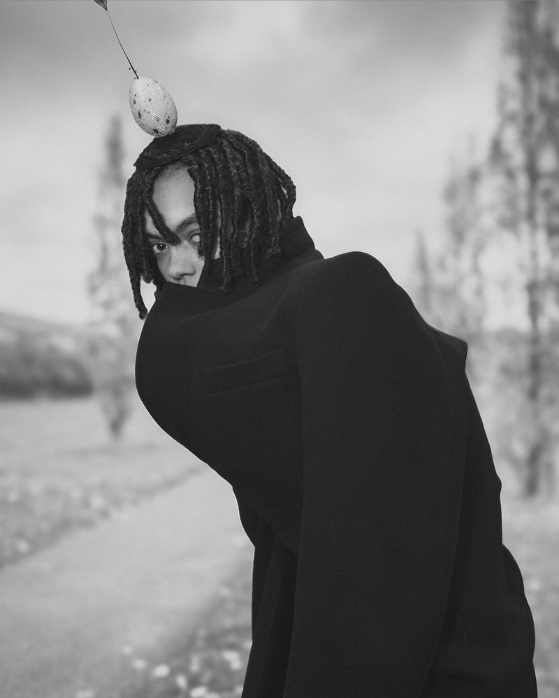 Portrait of person with dreadlocks and black clothing in a blurred outdoor setting, balancing a spotted egg on their head.