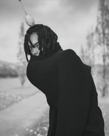 Portrait of person with dreadlocks and black clothing in a blurred outdoor setting, balancing a spotted egg on their head.