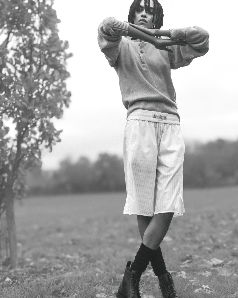 Monochrome portrait of a person with dreadlocks standing in a field, wearing casual clothes, boots, and posing with arms crossed.