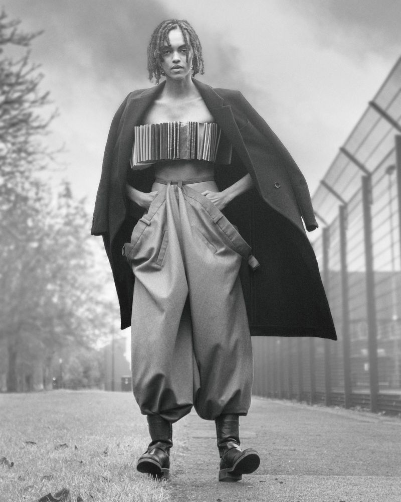 A woman in wide-leg pants and a coat poses on a path, wearing a book bodice, in a black and white fashion photograph against a cloudy sky.