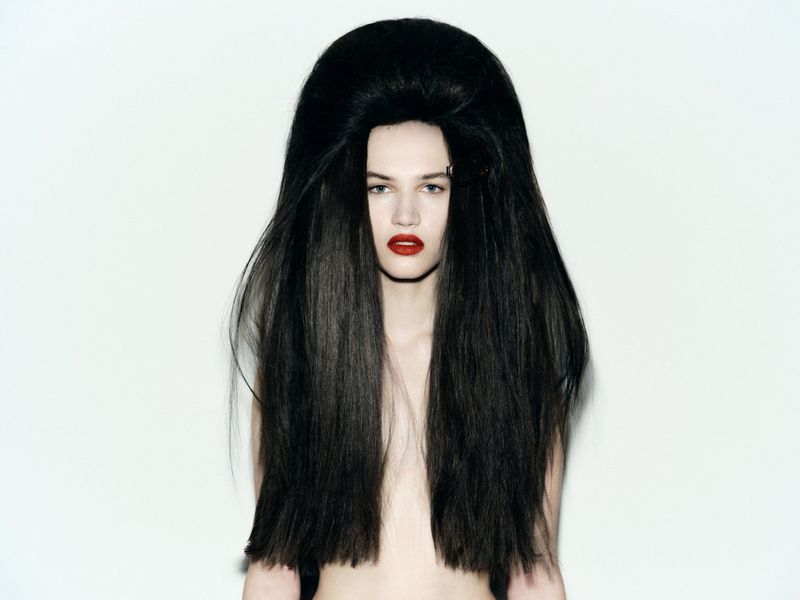 Striking portrait of a fair-skinned adult female model with a dramatic, oversized black bouffant hairstyle and bold red lipstick against white.