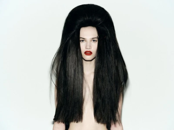 Striking portrait of a fair-skinned adult female model with a dramatic, oversized black bouffant hairstyle and bold red lipstick against white.