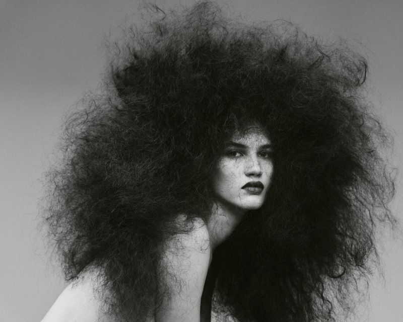 Monochrome portrait of a woman with dark hair styled in a large, voluminous afro, posing in front of a solid grey background.