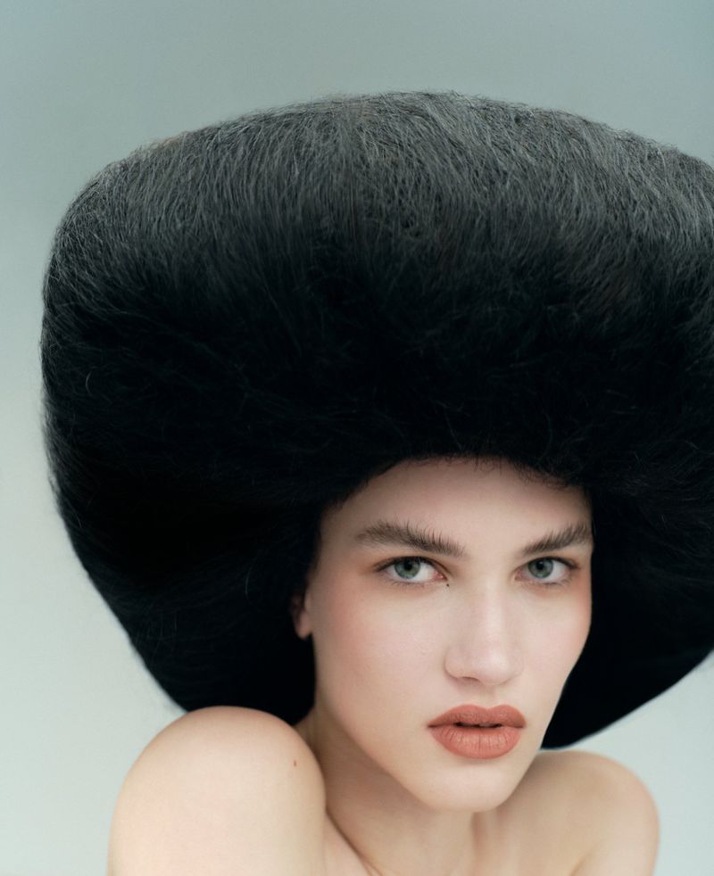 Glamour shot of a woman with bare shoulders and a striking black hairstyle against a pale blue background, fashion-forward.