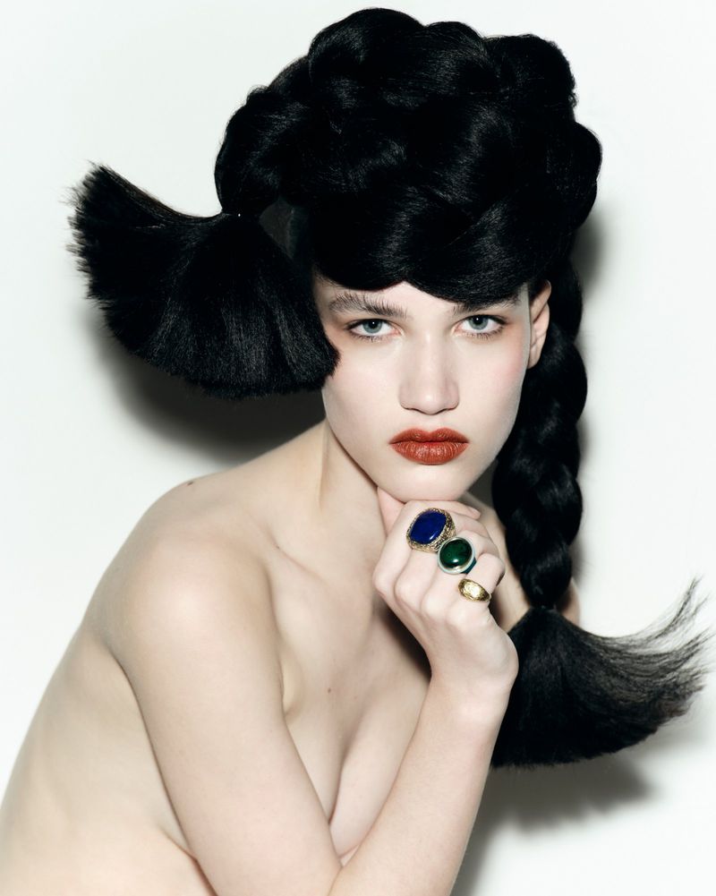 Portrait of a female model with dark hair styled in large braids, wearing rings, posing against a white background in a beauty shot.