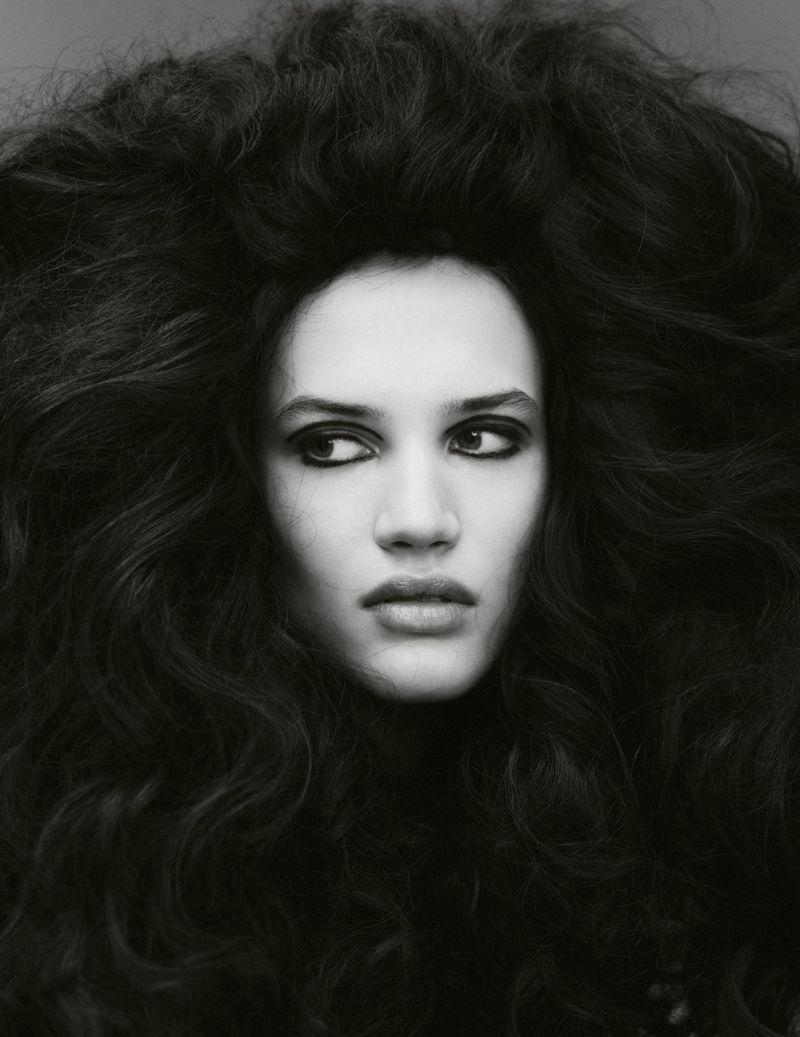 Monochrome portrait of a woman with voluminous dark wavy hair, gazing slightly to the side in a photo studio setting.