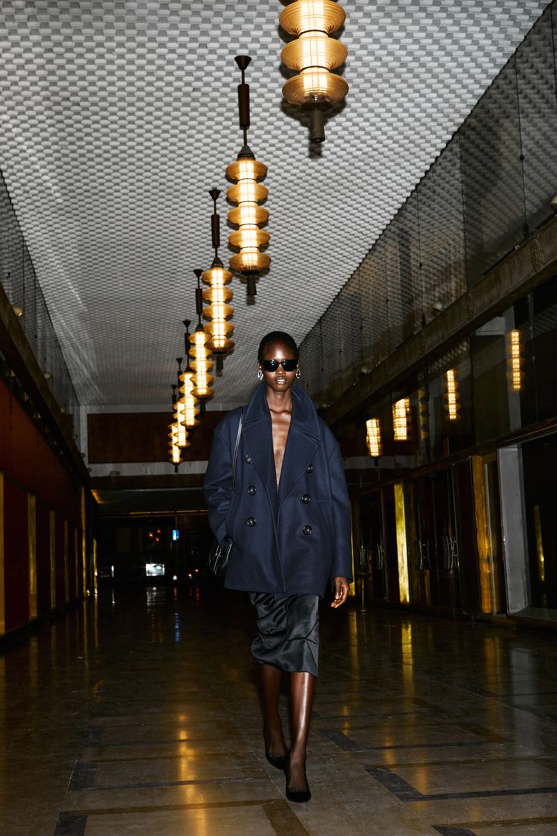 Fashionable adult female walks in a hallway wearing sunglasses, an open navy coat, a satin skirt, black shoes, and gold earrings.