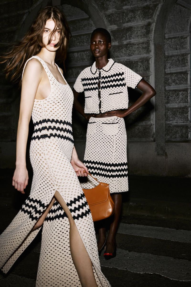 Two women in matching knitted dresses and high heels pose outside on a city street illuminated by streetlights at night, handbag.