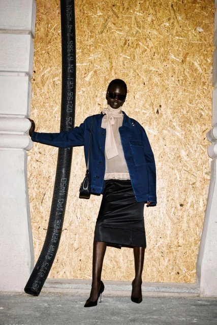 Fashion shot of an African American woman in a blue jacket and black skirt posing in front of a boarded up building with black pipe.