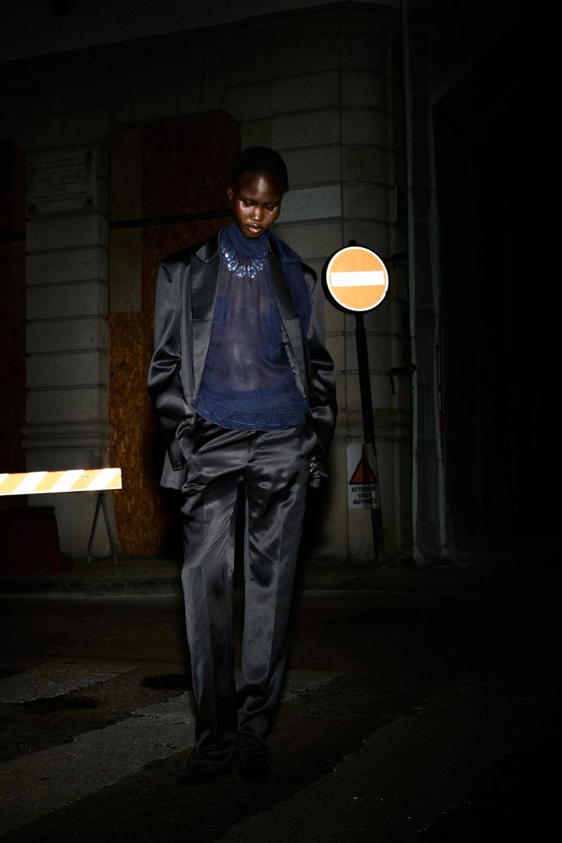 A slender Black model poses in a shiny dark pantsuit on a shadowy city street next to a traffic sign with a building in the background.