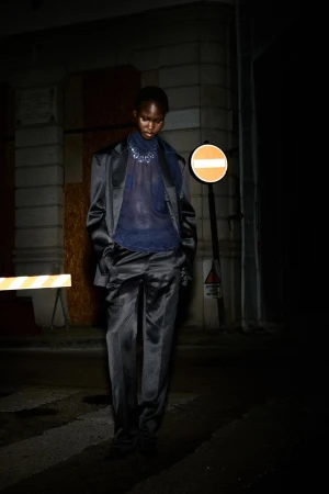 A slender Black model poses in a shiny dark pantsuit on a shadowy city street next to a traffic sign with a building in the background.