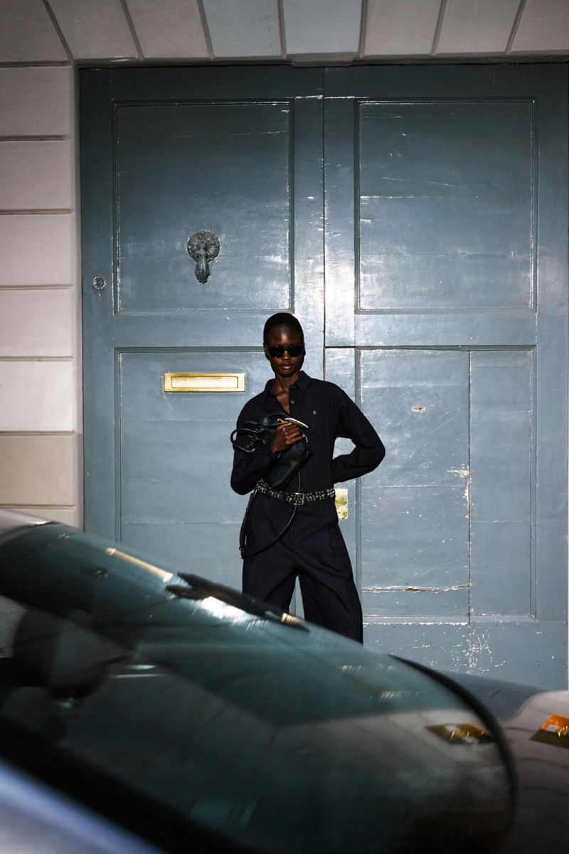 An adult female model of African descent in a dark jumpsuit and sunglasses poses in front of a blue door and partially obscured car.