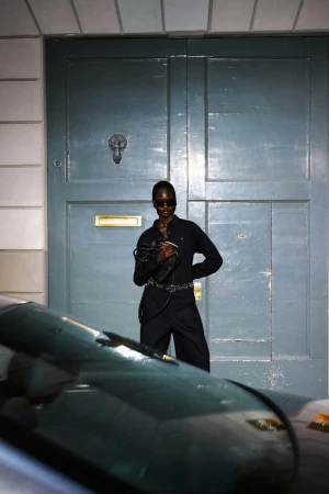 An adult female model of African descent in a dark jumpsuit and sunglasses poses in front of a blue door and partially obscured car.