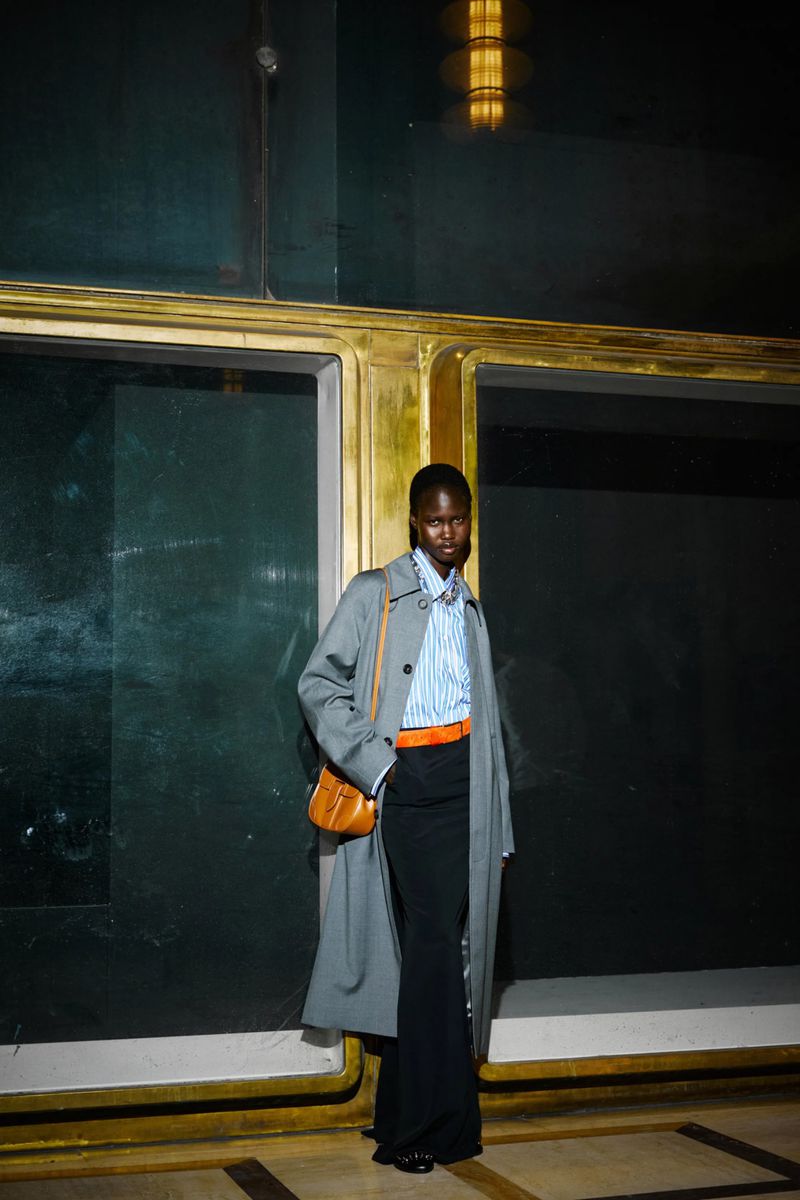A tall, dark-skinned woman is standing indoors wearing a trench coat, striped shirt, and long black skirt with a cross-body bag.